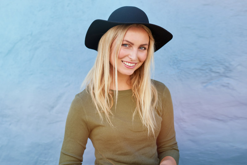 Nice lady with blonde hair and black hat smiling in front of a blue wall because she went to Wellington Family Dentistry & Implant Center for her tooth extraction
